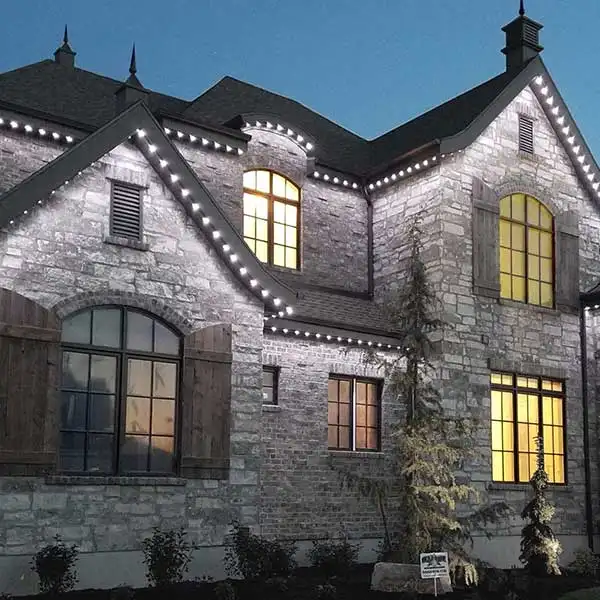 Stone two-story home at dusk with white permanent lighting outlining its rooflines and gables and warm light in the windows