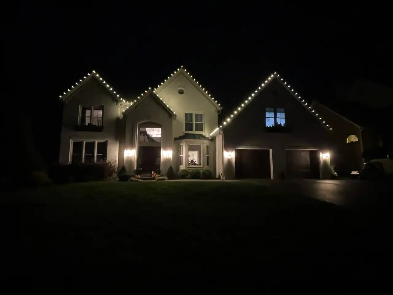 Two-story home in the Cincinnati area at night with warm white Christmas lights outlining three gables