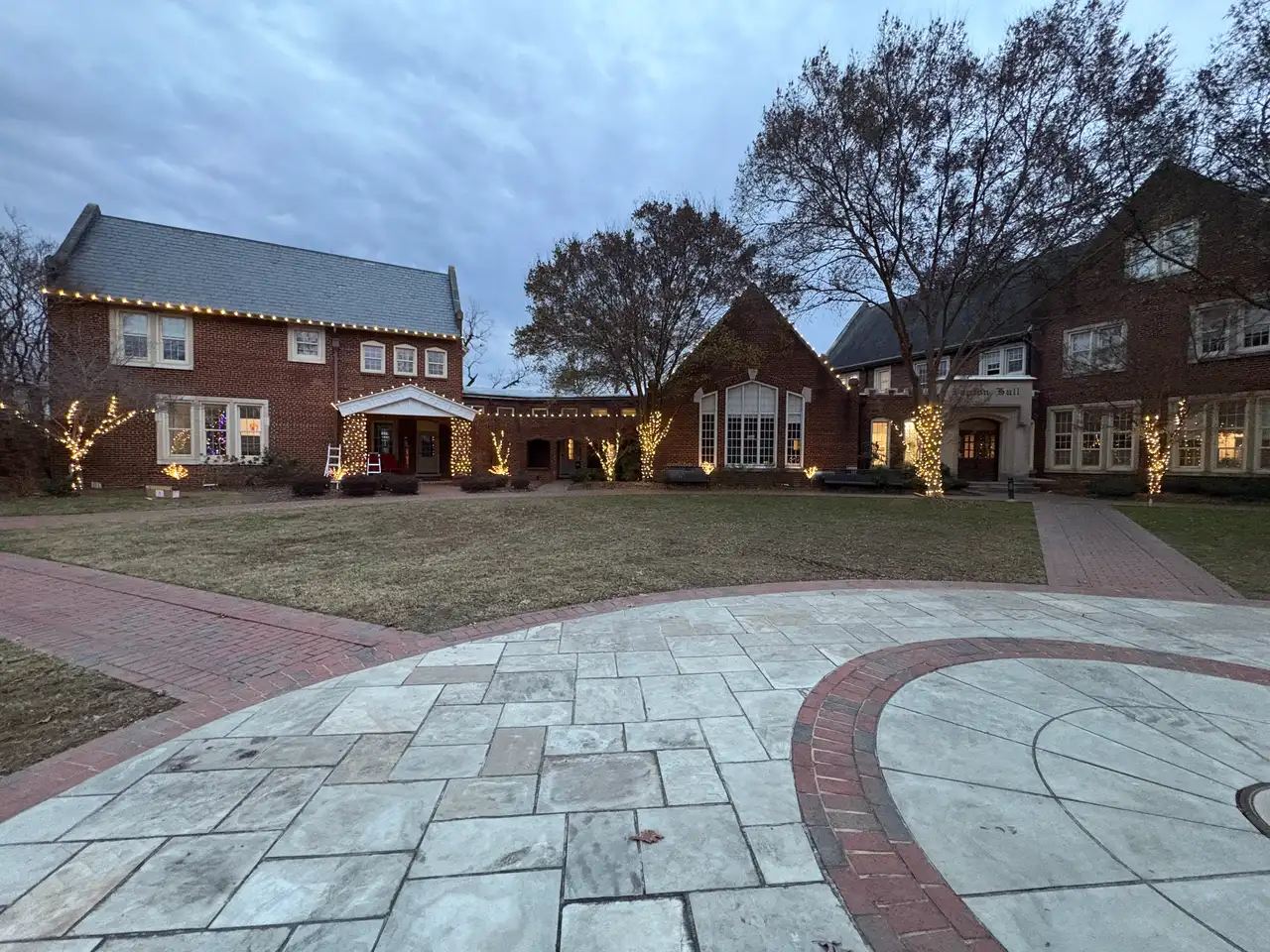 Brick buildings around a courtyard at Baylor School in Chattanooga at dusk with warm white holiday lights on the rooflines and wrapped trees