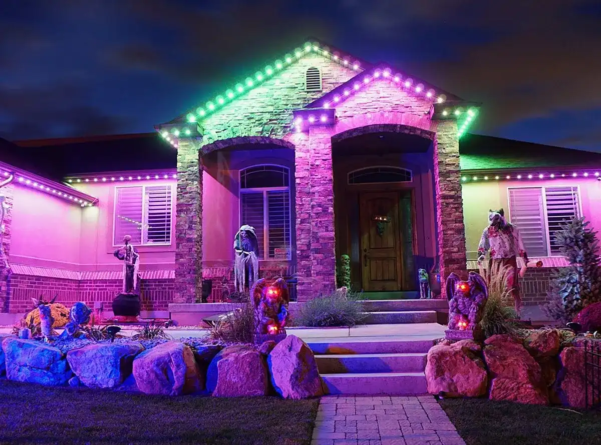 Stone-front home at night with green and purple permanent roofline lighting and Halloween figures by the front steps